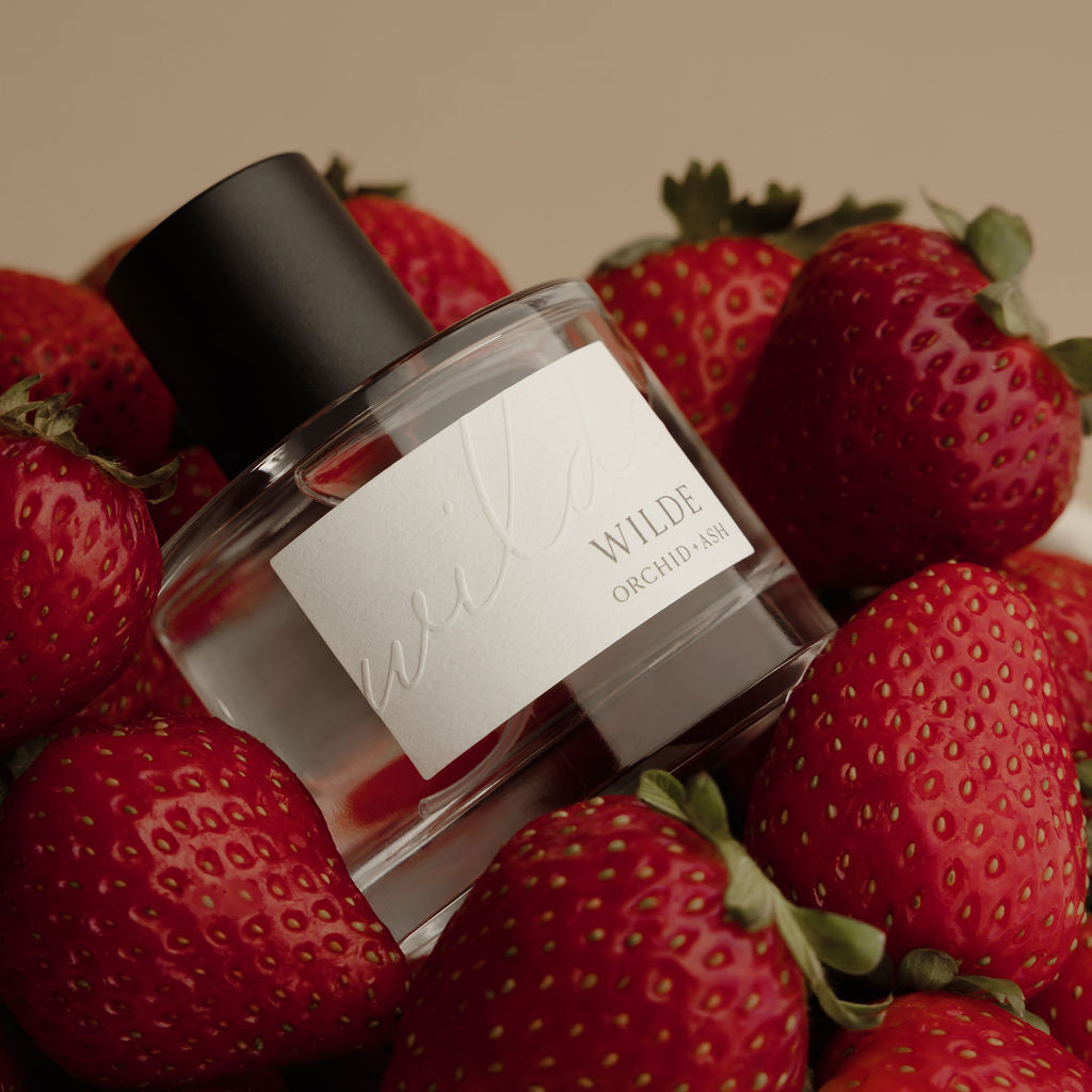 A clear glass perfume bottle with a black cap and a white label reading 'WILDE ORCHID' and 'MADISON SQUARE CITY', surrounded by fresh red strawberries against a warm beige background.