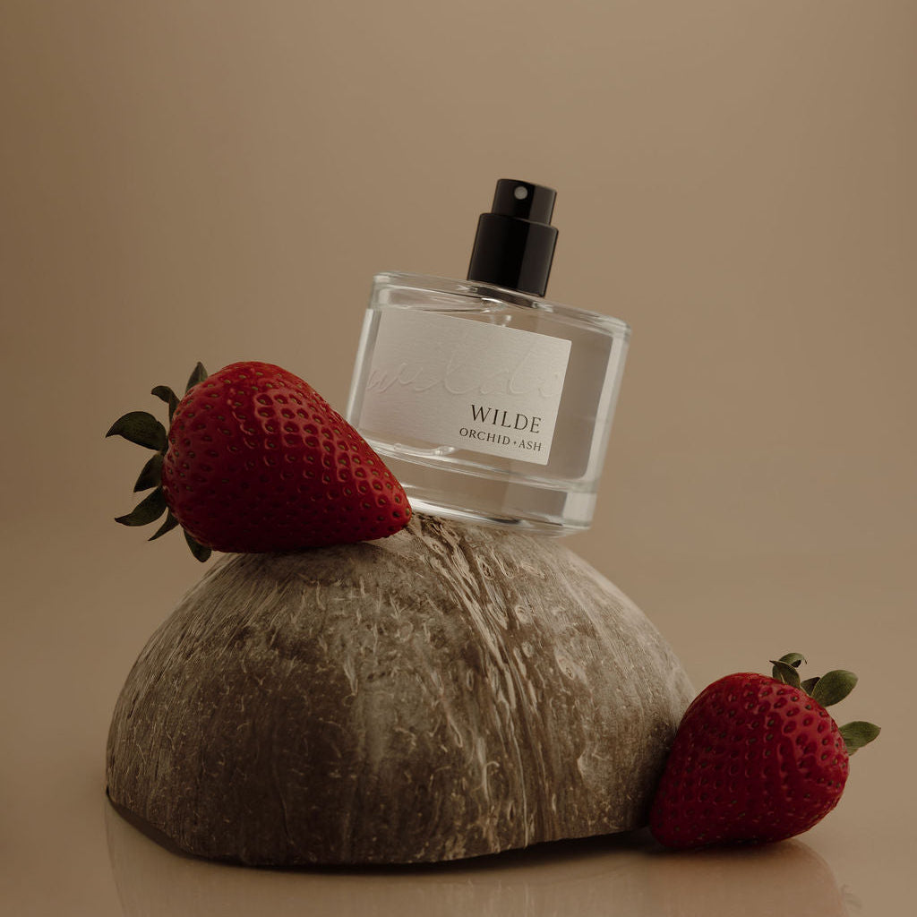 A clear rectangular perfume bottle with a black spray top and a white label reading WILDE, styled on a textured stone slab with two red strawberries on a warm beige background.