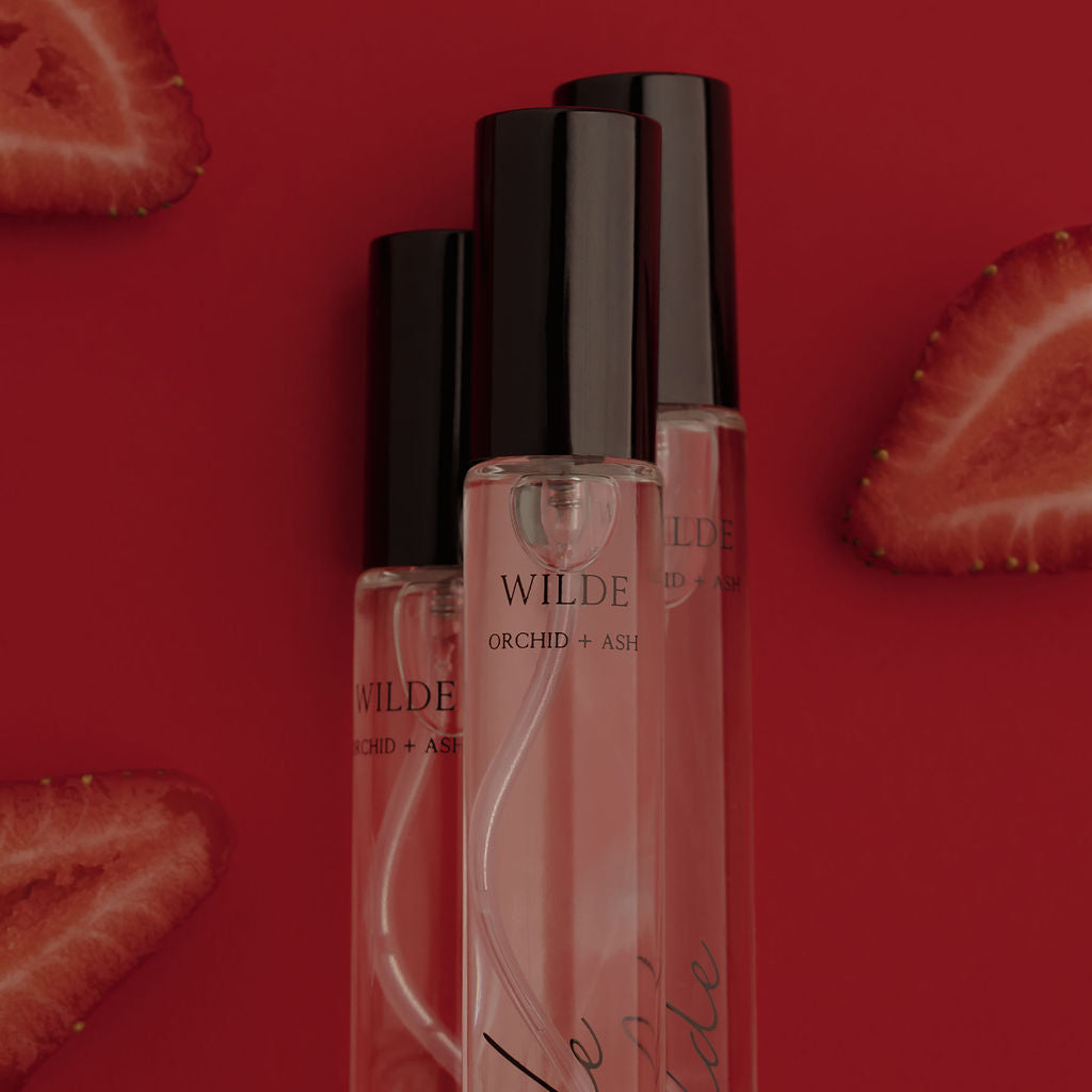 Three clear cosmetic bottles with black caps and pink liquid, labeled WILDE Orchid + Air, arranged on a red background with strawberry slices around them.