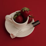 A clear glass perfume roller bottle with a black cap and the brand name 'wilde' printed on the tube, styled on a white saucer with a white teacup filled with fresh red strawberries, all set against a solid deep red background.