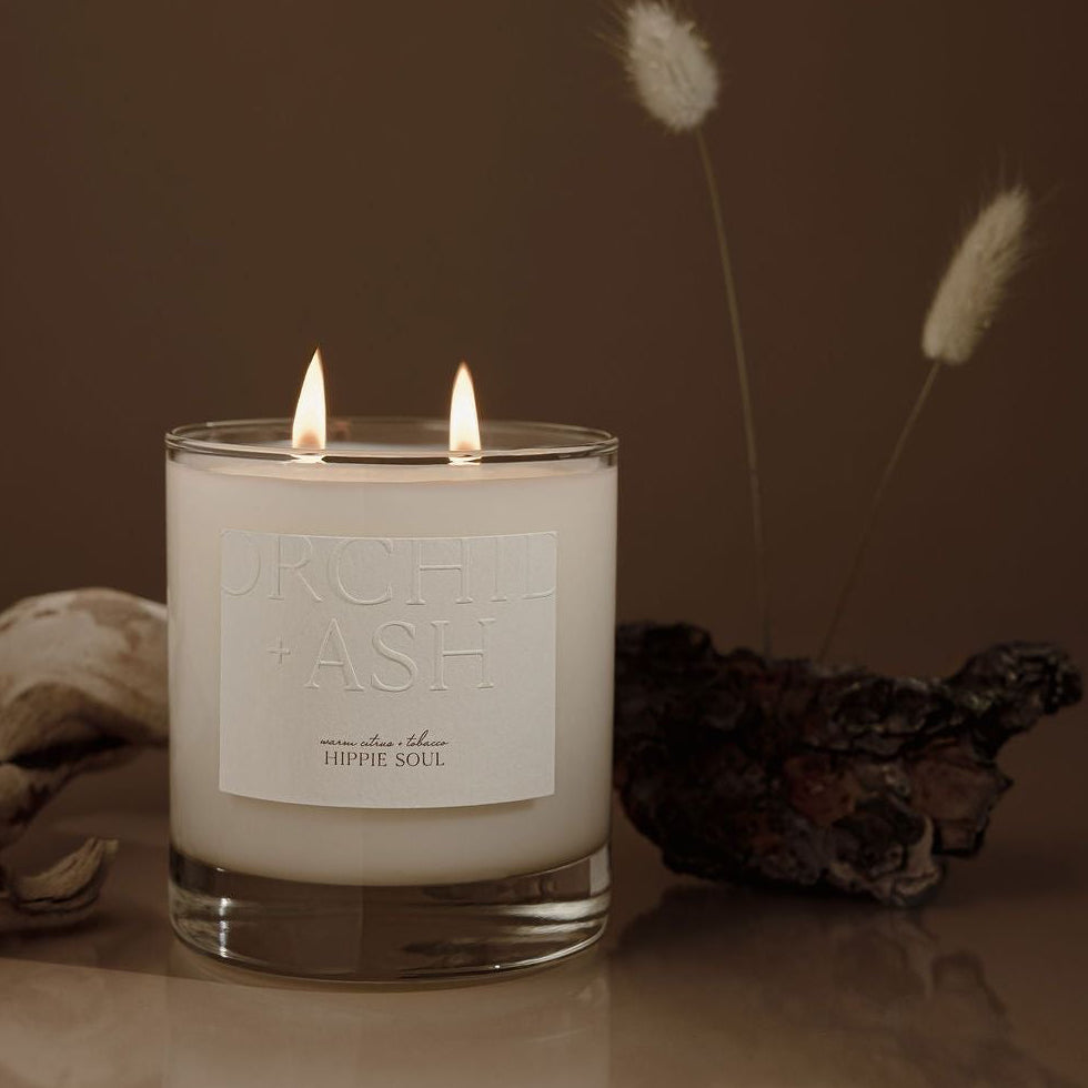 Candle in a glass jar with a label on a reflective surface, surrounded by dried plants.
