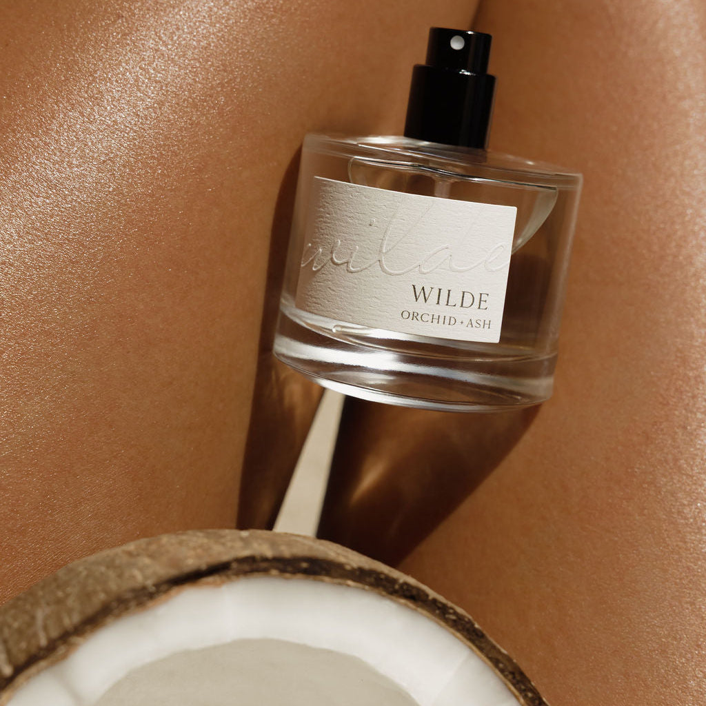 A clear glass perfume bottle labeled 'WILDE ORCHID JAZZ' with a black spray top, styled on a warm beige skin-toned surface with a cut coconut in the foreground.