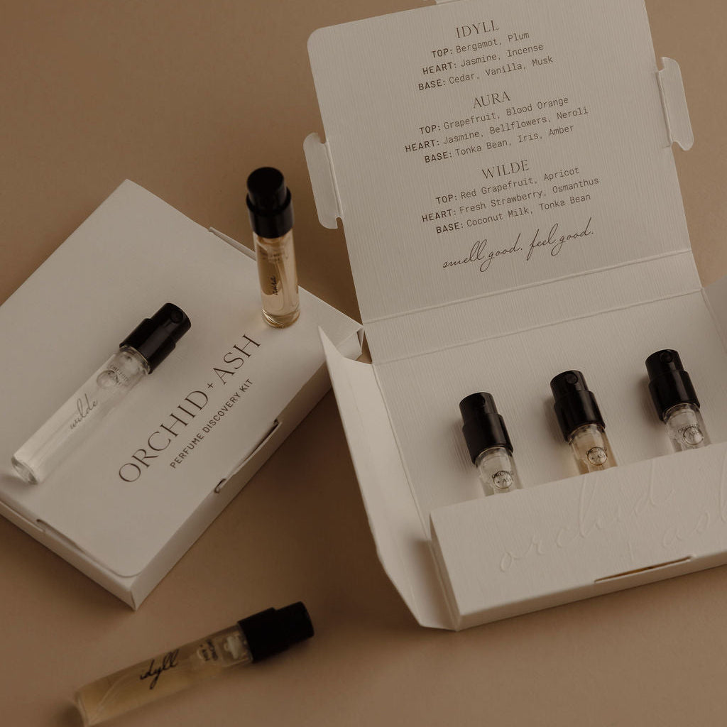 A set of white perfume sample vials and boxes from Orchid Ash, arranged on a warm beige background. One open box displays four clear glass vials with black caps, while another box lies beside it with a single vial resting on top.