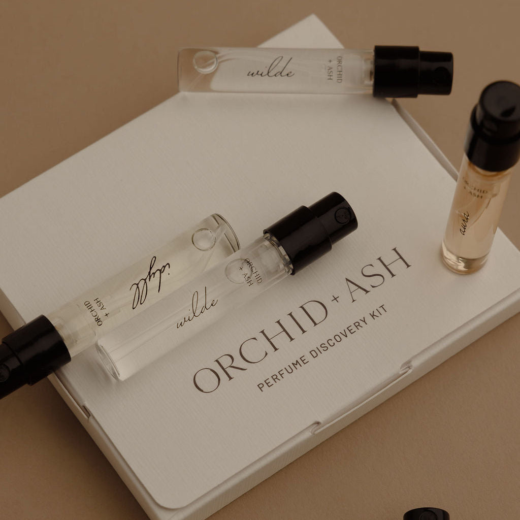 Orchid & Ash Perfume Discovery Kit featuring a white box with several clear glass sample spray vials and one small amber bottle, arranged on a warm beige background.