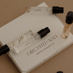 Orchid & Ash Perfume Discovery Kit featuring a white box with several clear glass sample spray vials and one small amber bottle, arranged on a warm beige background.