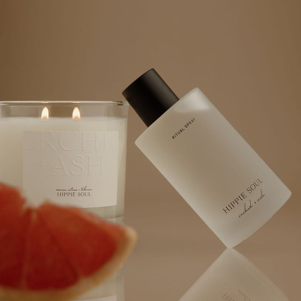 A frosted glass bottle labeled HIPID SOUL and a lit candle in a glass jar, styled with a grapefruit slice in the foreground against a warm beige background.