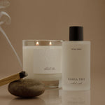 A Joshua Tree scented candle in a clear glass jar with two lit flames and a white bottle with a black cap beside it, styled on a reflective surface with a stone and a smoking incense stick against a warm beige background.