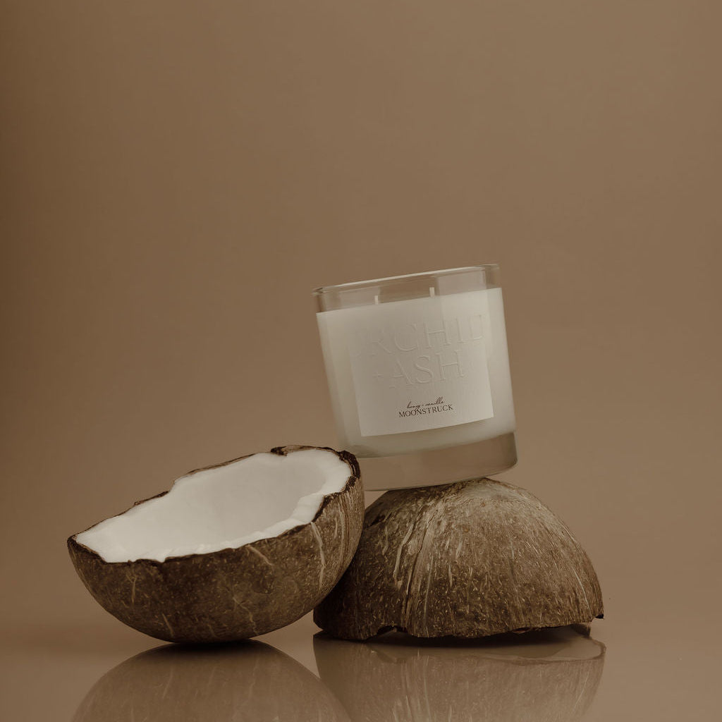 Candle in a glass container with coconut halves on a reflective surface against a brown background