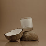 Candle in a glass container with coconut halves on a reflective surface against a brown background