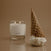 Candle in a glass container next to an ice cream cone on a reflective surface with a beige background