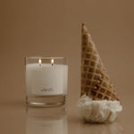 Candle in a glass container next to an ice cream cone on a reflective surface with a beige background