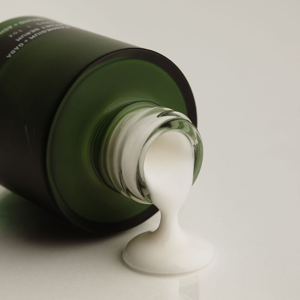 A dark green cosmetic bottle lying on its side with a thick white cream or lotion dripping from the open neck onto a clean white surface.