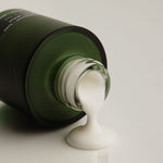 A dark green cosmetic bottle lying on its side with a thick white cream or lotion dripping from the open neck onto a clean white surface.