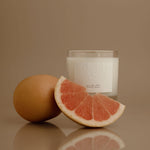 Candle with a grapefruit slice and whole grapefruit on a reflective surface against a brown background