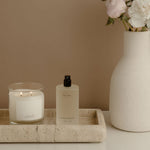 Candle, bottle, and vase with flowers on a marble tray against a beige wall.