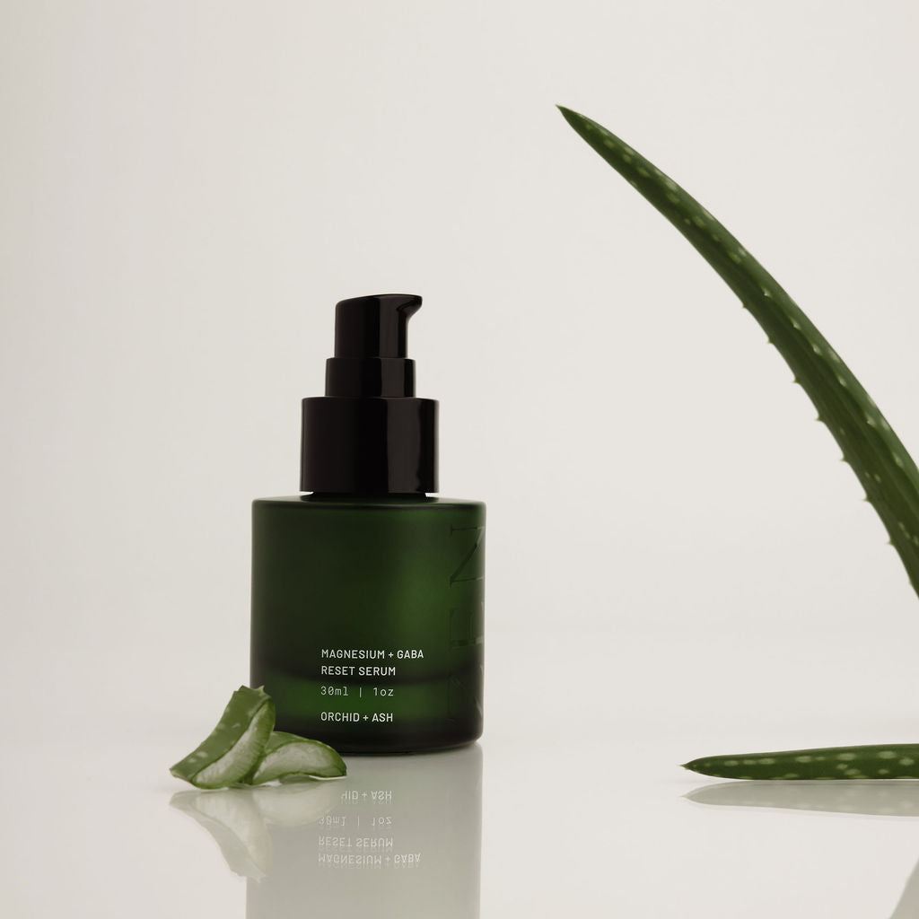 A dark green pump bottle of ORCHE + AEP Fruit Serum with a black pump, styled on a reflective white surface with aloe vera slices and a long aloe leaf in the background.