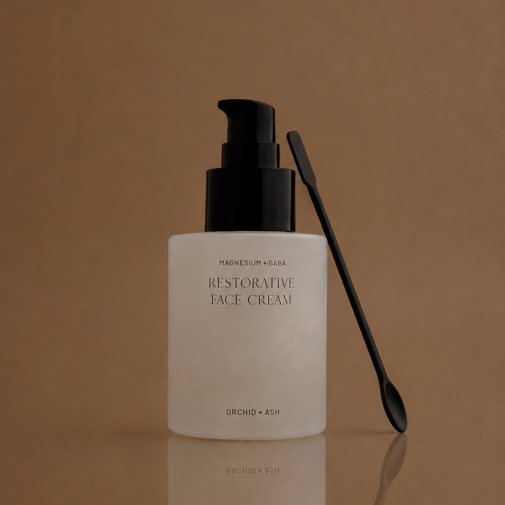 A frosted white bottle of Magnesium & Kiehl Restorative Face Cream with a black pump, shown next to a black spatula on a reflective brown surface.
