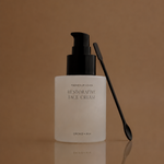 A frosted white bottle of Magnesium & Kiehl Restorative Face Cream with a black pump, shown next to a black spatula on a reflective brown surface.