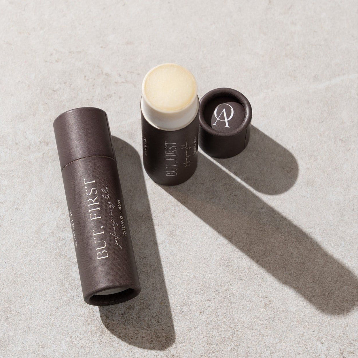 Three cosmetic products on a textured surface with 'BUT. FIRST' branding.