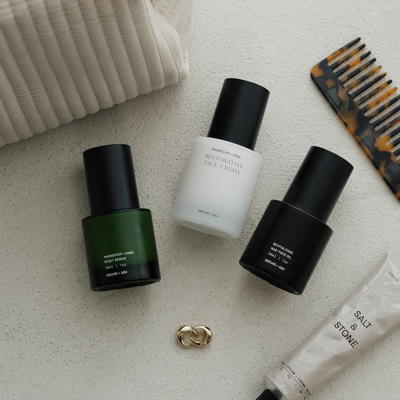 Skincare products on a textured surface with a comb and jewelry.