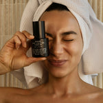 Person holding a bottle of face oil with a towel on their head, against a neutral background